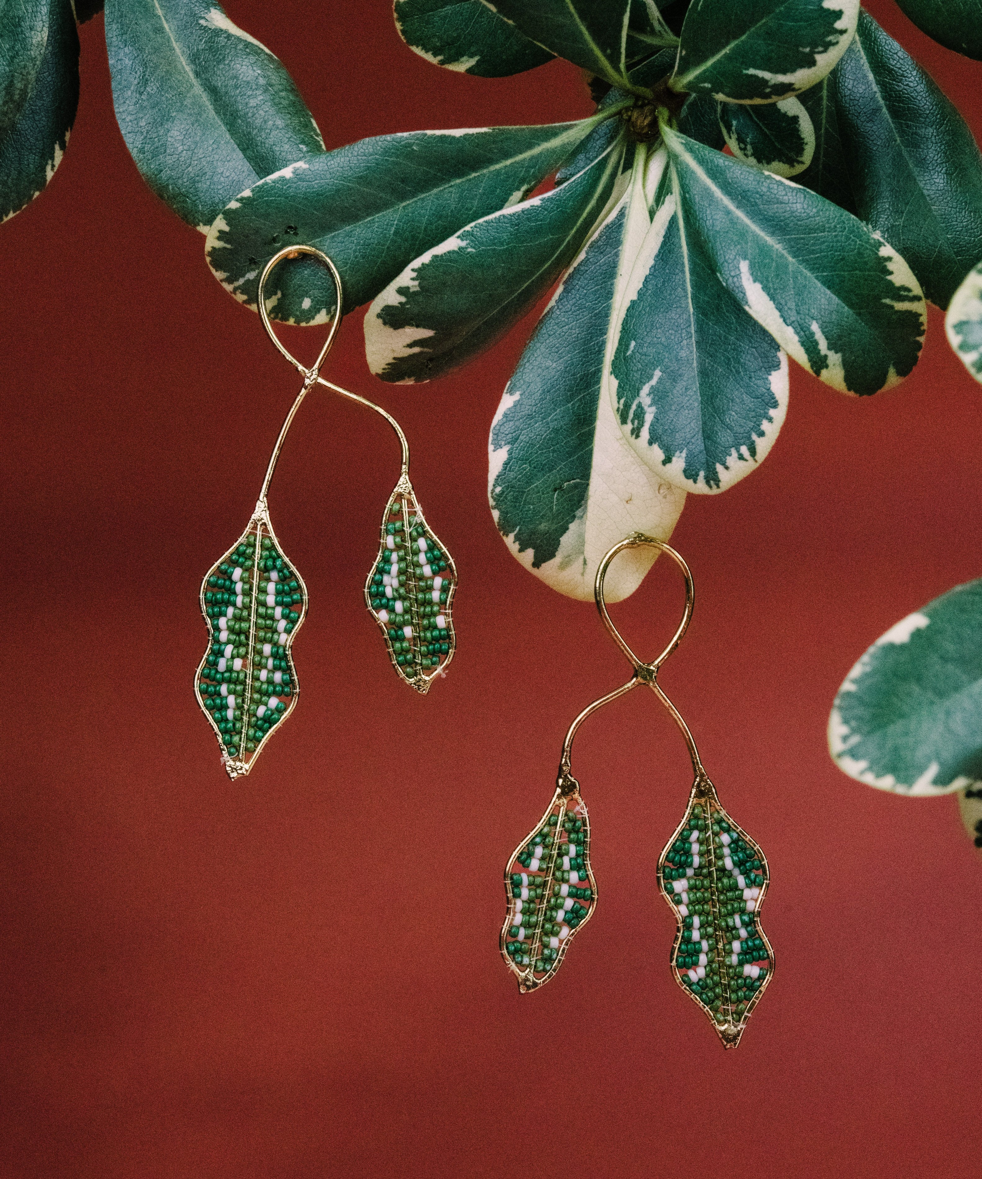 Green leaf-shaped earrings with embedded stones against a red background with leaves.