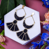 Black and gold earrings on a white hexagonal stand with flowers and leaves on a pink background