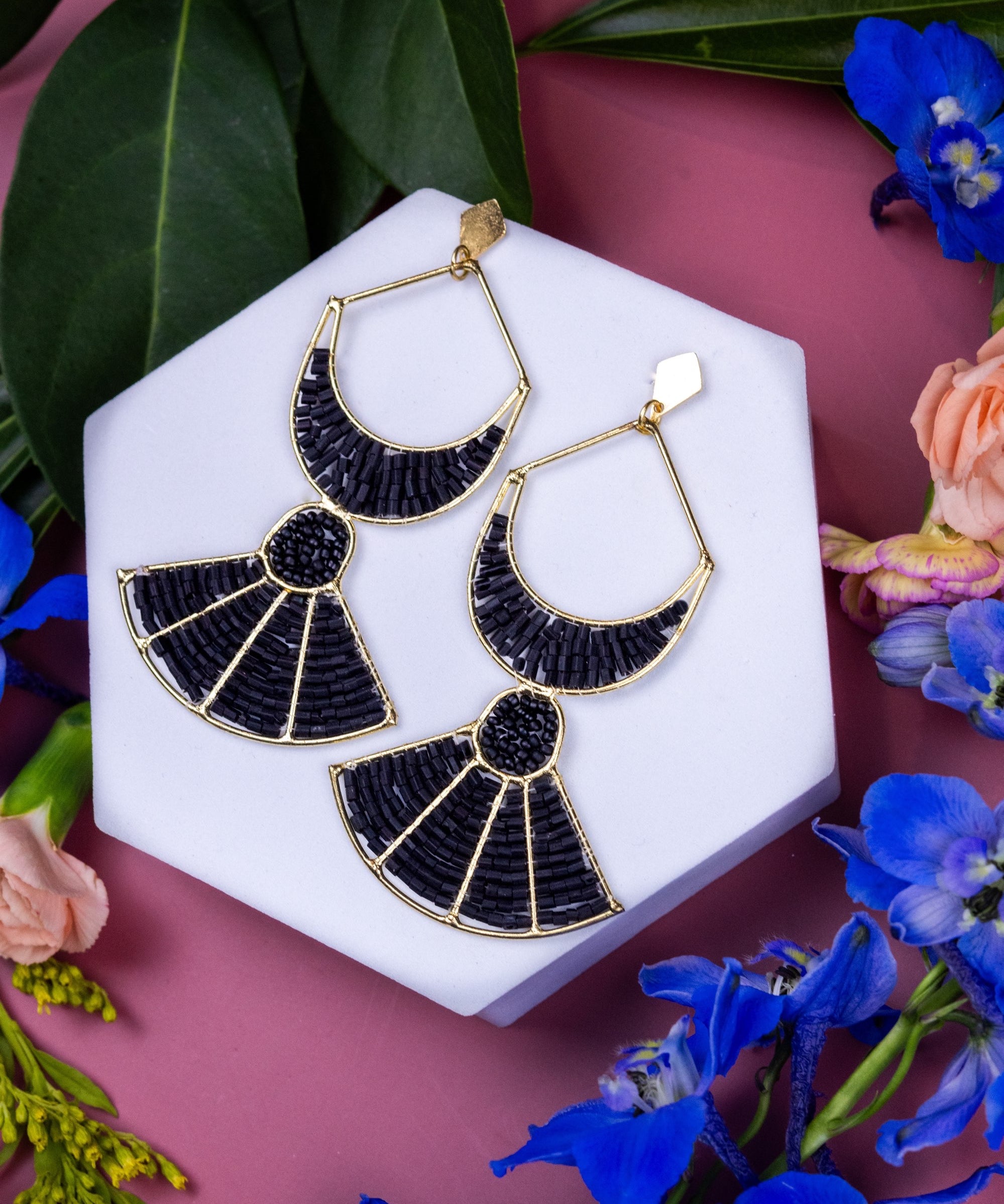 Black and gold earrings on a white hexagonal stand with flowers and leaves on a pink background