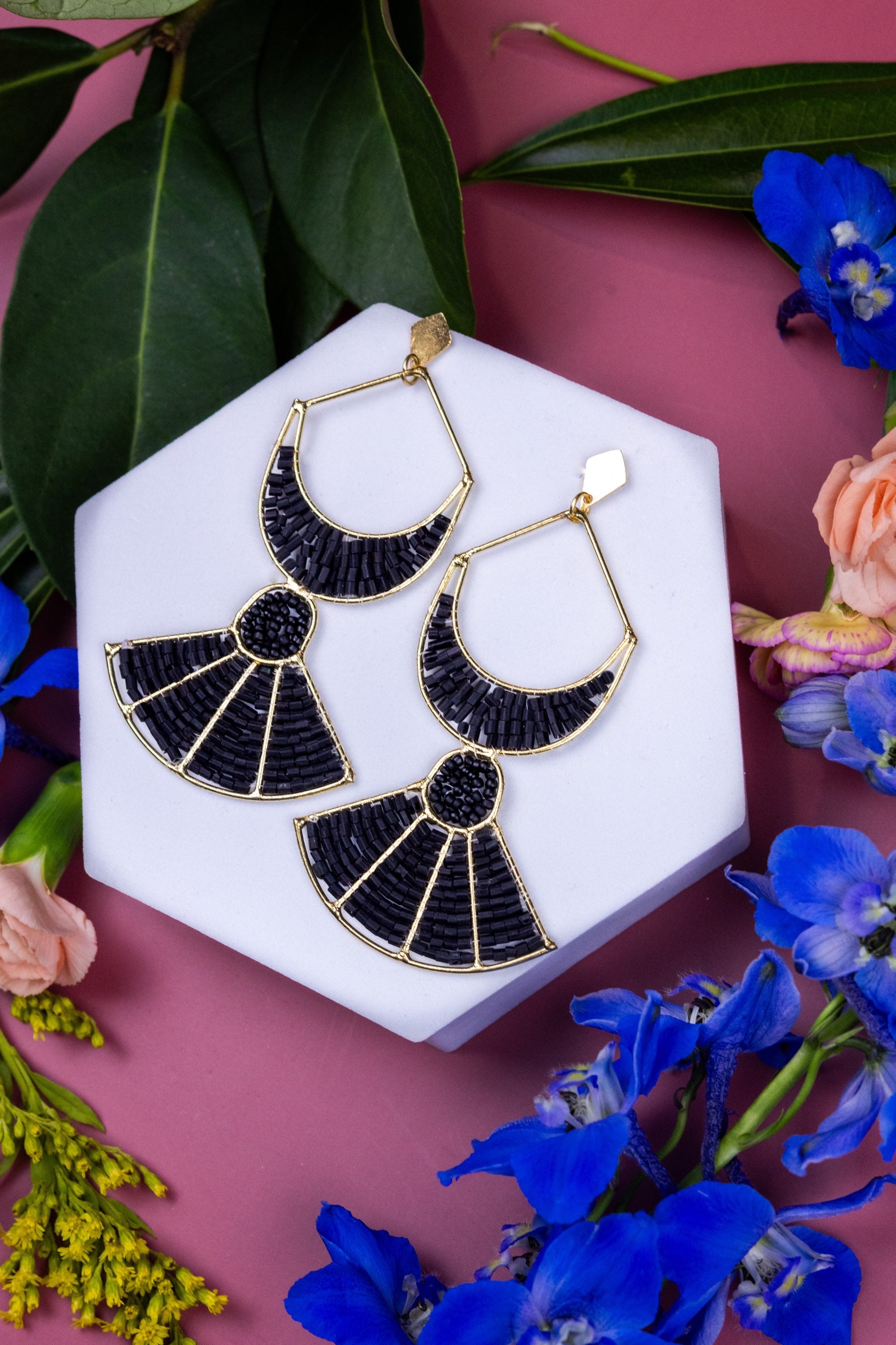 Black and gold earrings on a white hexagonal stand with flowers and leaves on a pink background