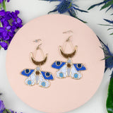 Pair of blue and gold earrings on a pink round surface with purple flowers and green leaves.