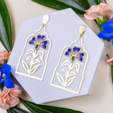 Gold earrings with blue wildflower design on a gray card with green leaves and flowers in the background