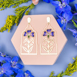 Decorative earrings on a hexagonal pink stand with flowers around on a blue background