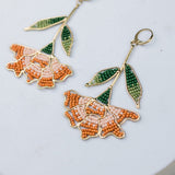 Pair of floral earrings with orange flowers and green leaves on a light blue circular stand.