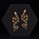 Snake-shaped earrings with red, black, and gold details on a black hexagonal stand.