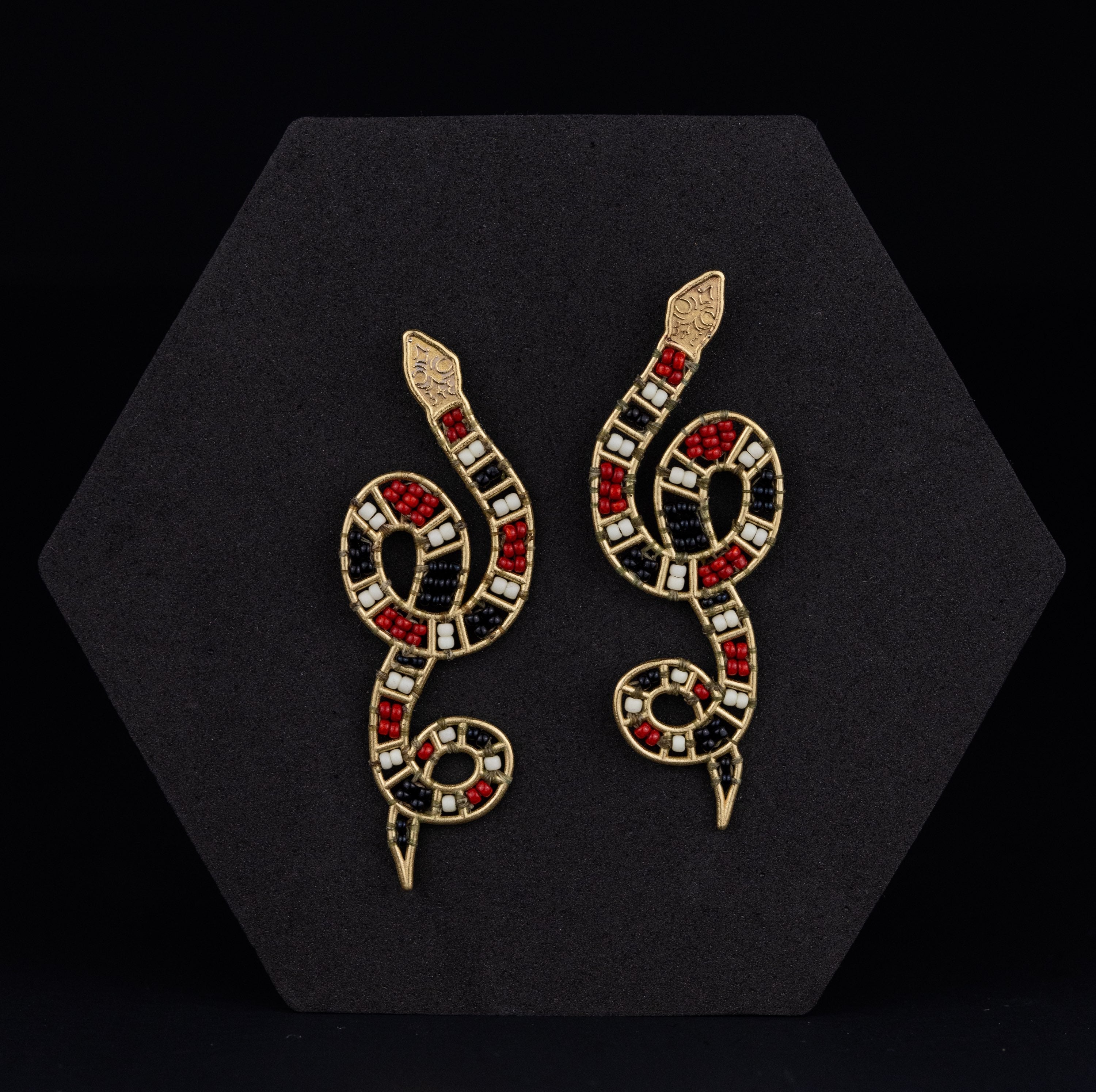 Snake-shaped earrings with red, black, and gold details on a black hexagonal stand.
