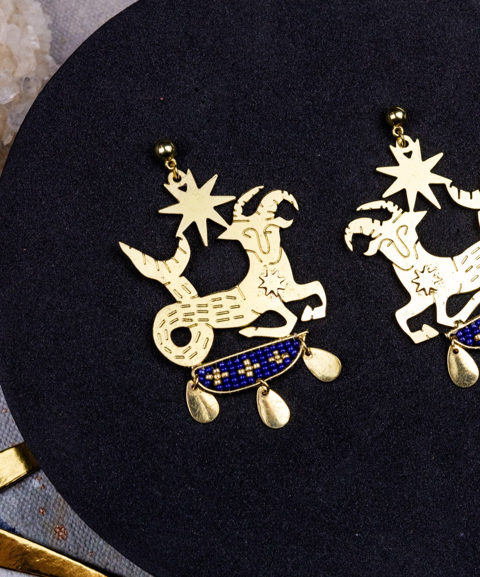 Gold zodiac earrings on a black circular stand with a textured surface.