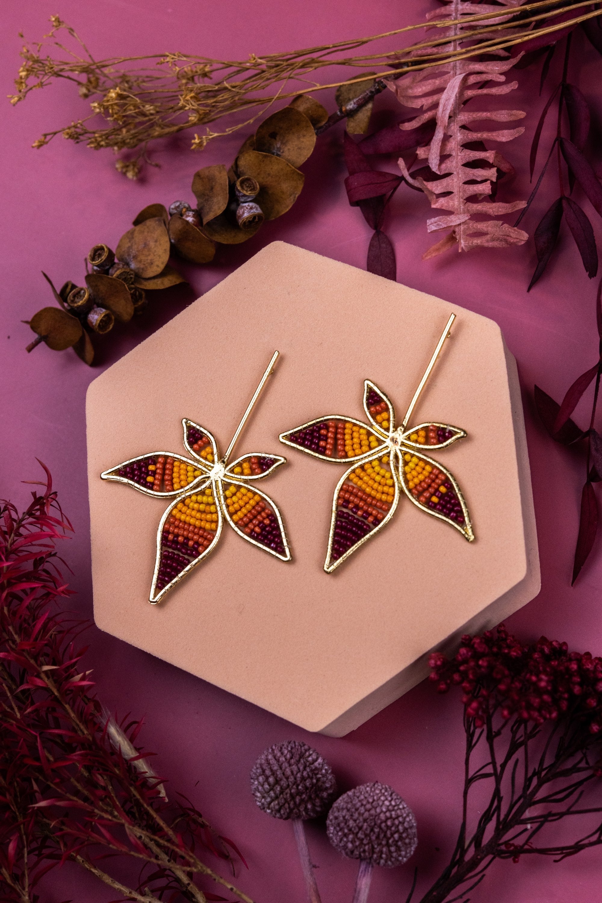 Leaf-shaped earrings on a hexagonal pink stand with dried leaves and flowers on a pink background