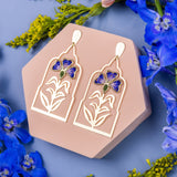 Gold earrings with butterfly design on a hexagonal pink stand surrounded by blue flowers.