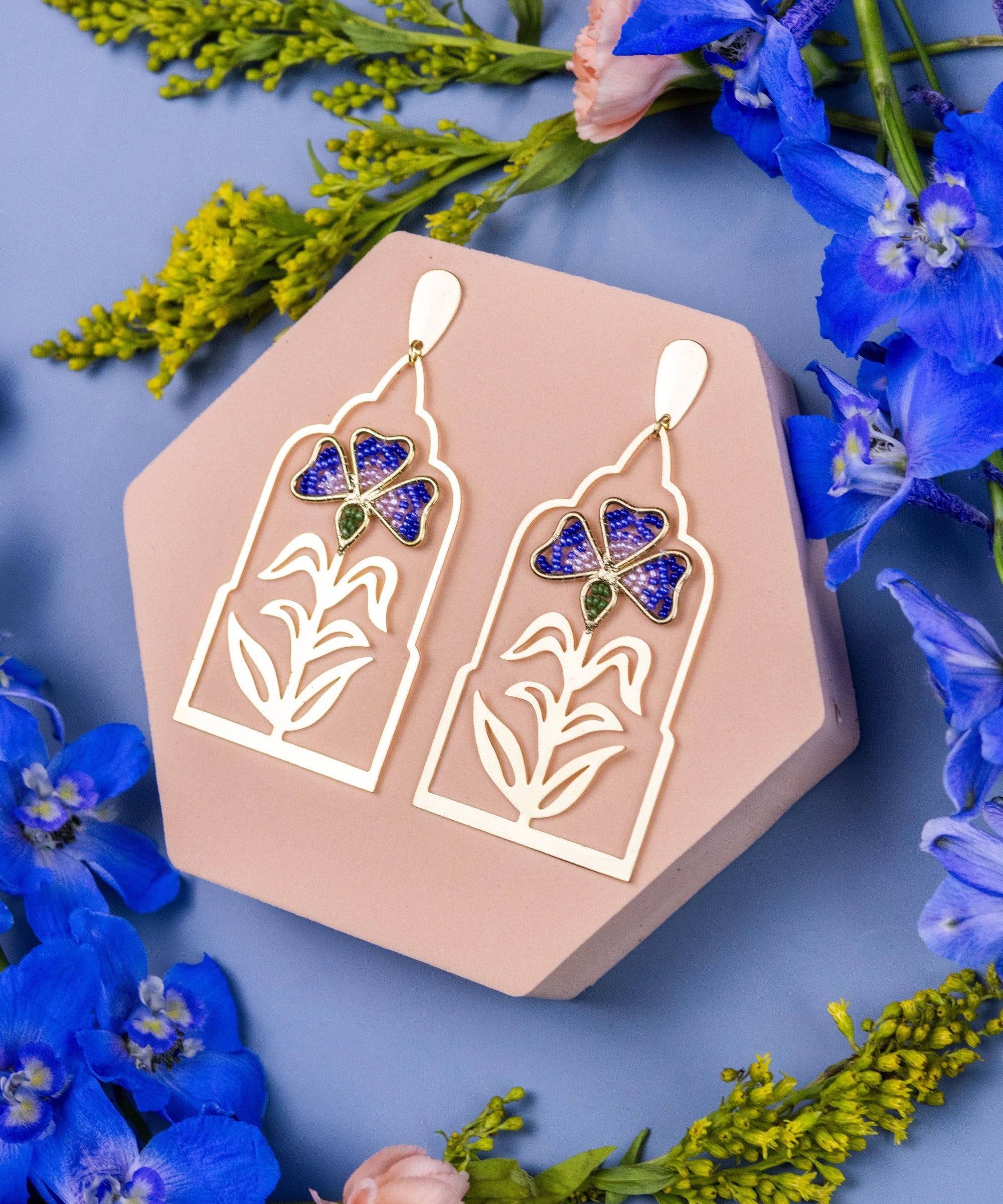 Gold earrings with butterfly design on a hexagonal pink stand surrounded by blue flowers.