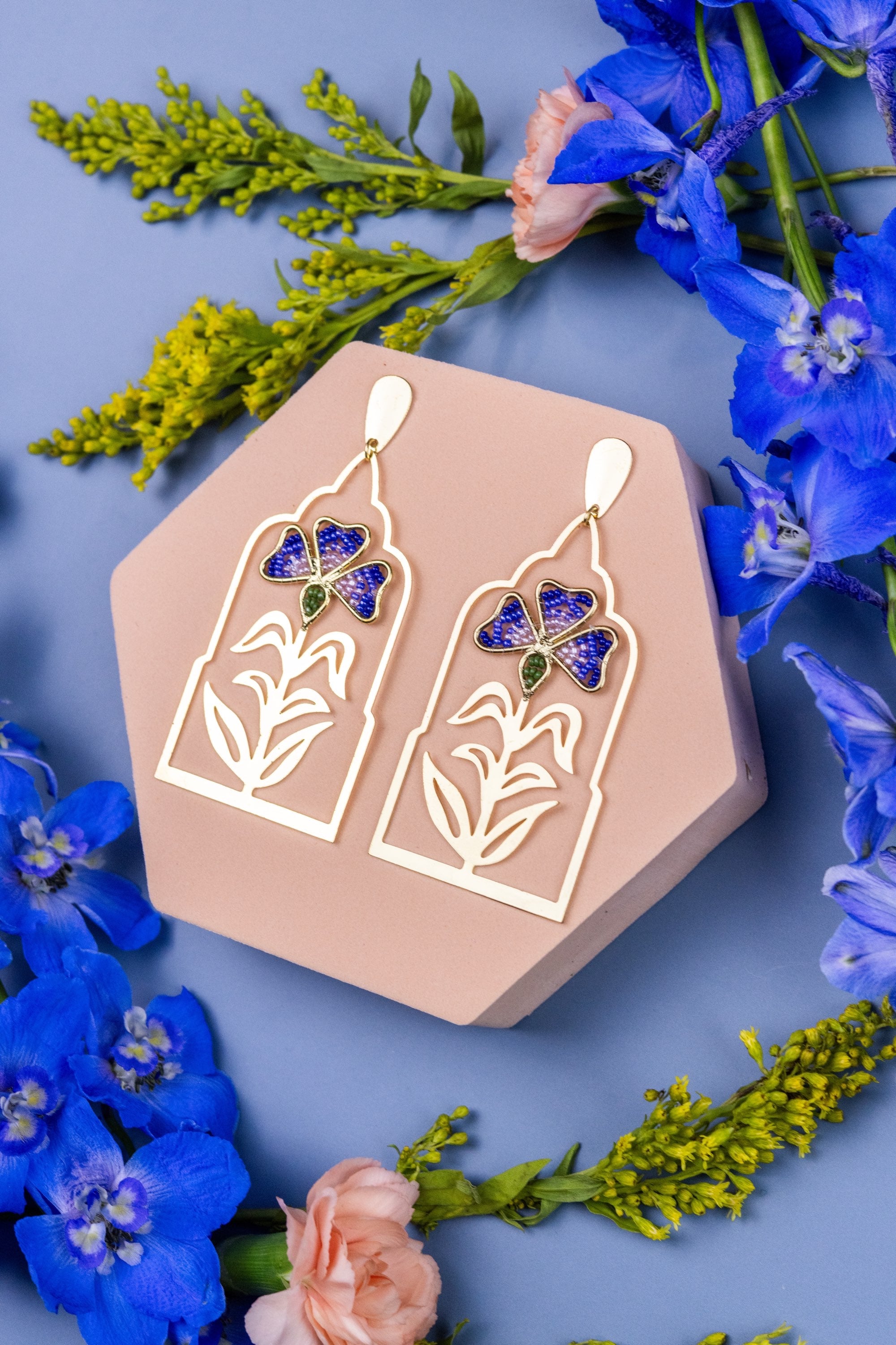 Gold earrings with butterfly design on a hexagonal pink stand surrounded by blue flowers.